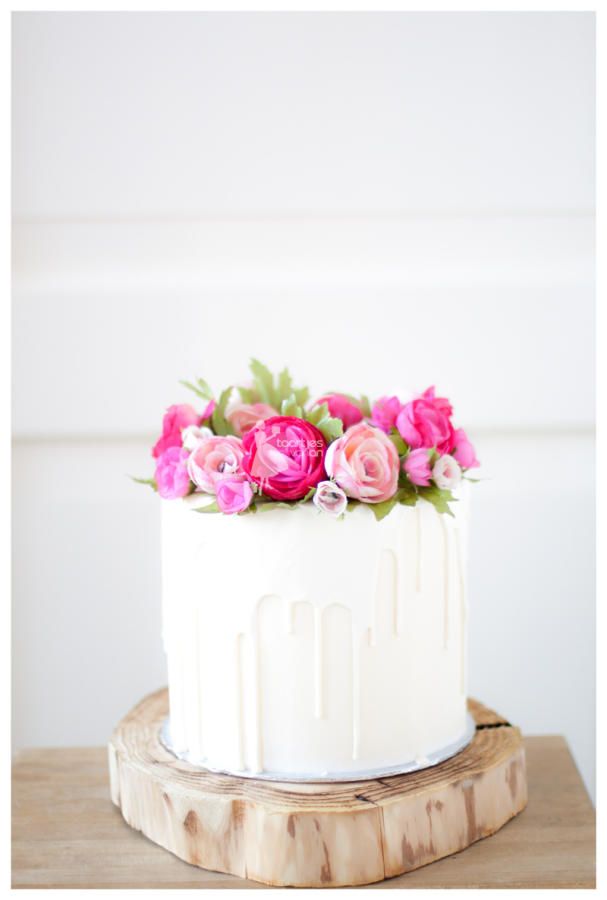 a white cake topped with pink flowers on top of a wooden stand next to a wall