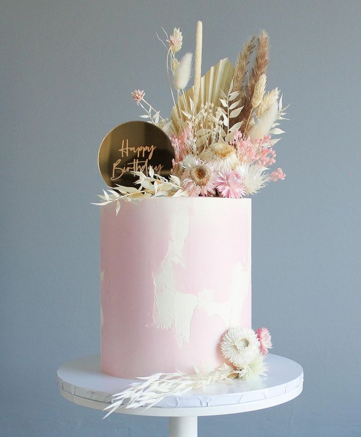 a pink cake with flowers and feathers on top is sitting on a white stand against a gray wall