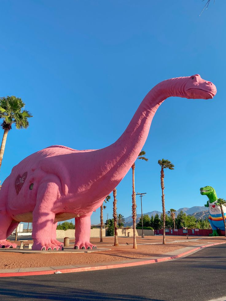 an inflatable pink dinosaur is on the side of the road near palm trees