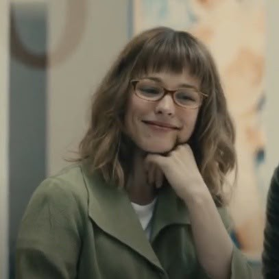 a woman with glasses sitting in front of a mirror looking at the camera and smiling