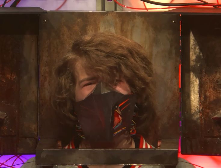 a man wearing a black face mask on top of a tv screen with red and purple lights behind him