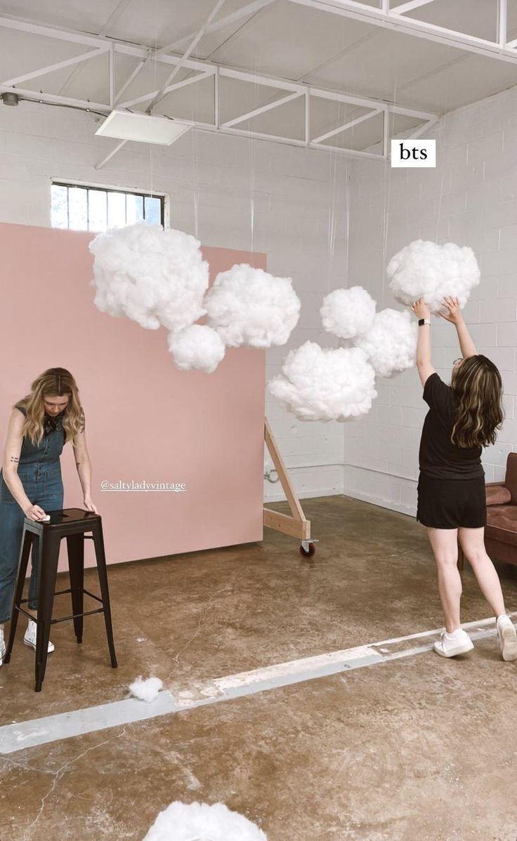 two women standing in front of a pink wall with white clouds floating over them and one woman sitting on a stool