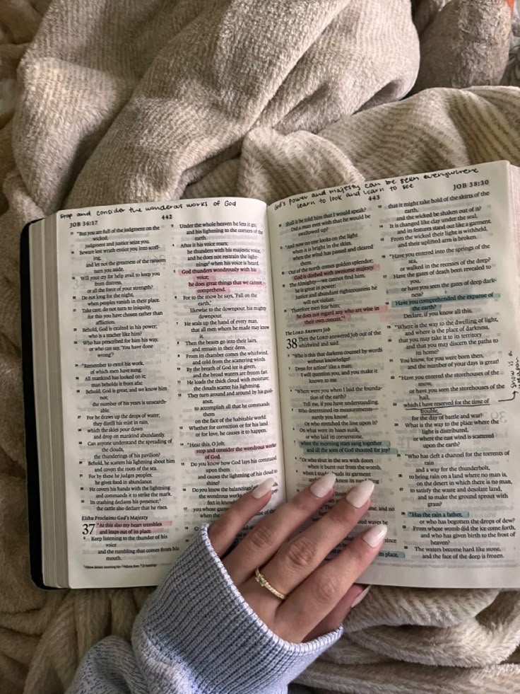 a woman is holding an open book in her hands while laying on a bed covered with blankets