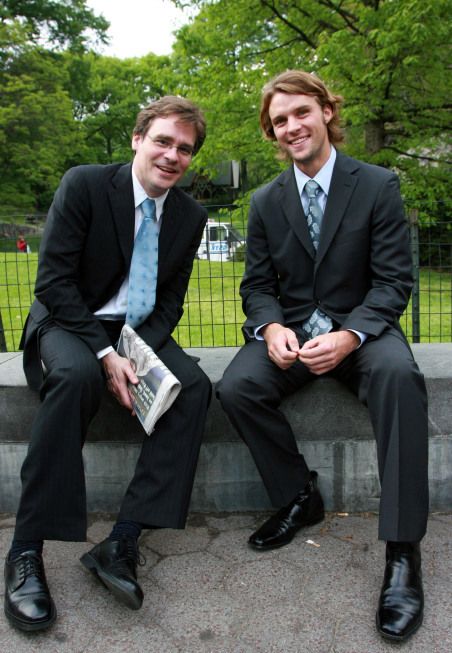 two men in suits and ties sitting next to each other on a cement bench with trees in the background