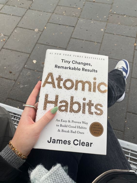 a person holding up a book about atomic habitts in front of their feet on the sidewalk