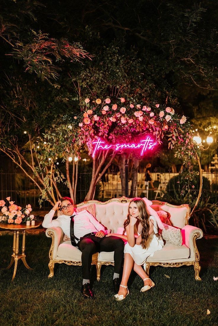 a man and woman sitting on a couch under a pink neon sign