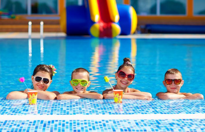 four children in sunglasses are sitting on the edge of a swimming pool and drinking cocktails