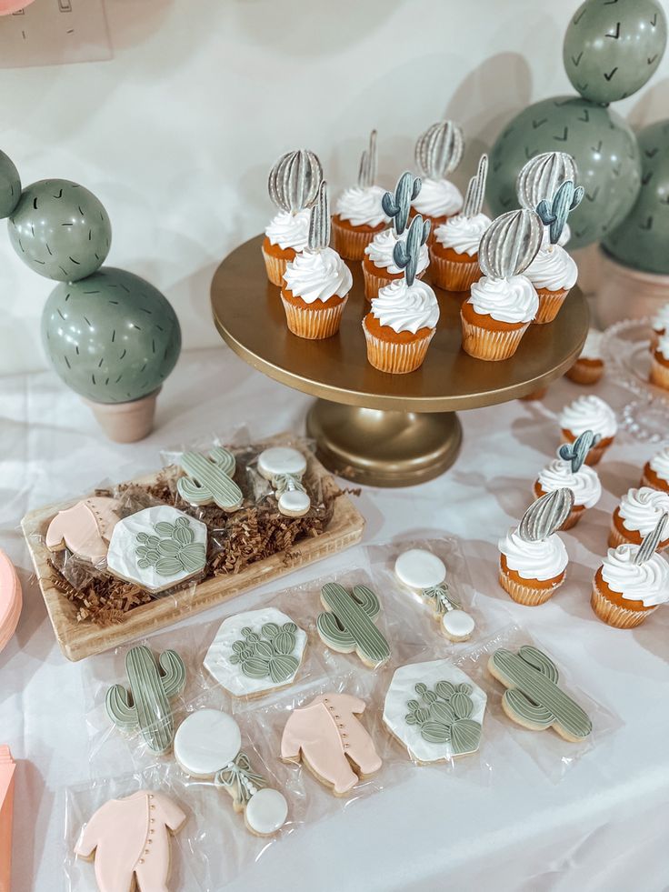 desserts and cupcakes are displayed on a table with cactus themed decorations in pastel colors