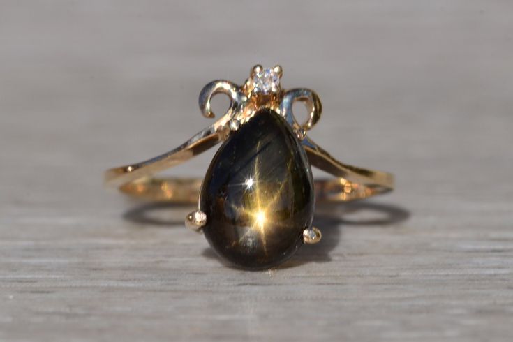 The Towner Bay: Ladies Vintage Black Star Sapphire Ring in Yellow Gold. Introducing the stunning and luxurious yellow gold ring with a magnificent 3 carat pear-shaped black star sapphire as its centerpiece. The ring features a beautiful 3 carat black star sapphire that has been carefully selected for its unique and mesmerizing beauty. The sapphire's pear shape adds a touch of elegance to the piece and is sure to be the envy of any collection. To further enhance its allure, a sparkling diamond ac Black Star Sapphire Engagement Ring, Vintage Black Ring, Elegant Star-shaped Sapphire Ring For Anniversary, Elegant Star-shaped Sapphire Ring, Black Star Sapphire Ring, Black Diamond Engagement Ring Vintage, Star Sapphire Engagement Ring, Black Sapphire Ring, Black Star Sapphire