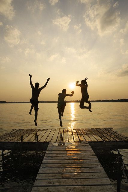 three people jumping into the air from a dock at sunset or dawn, with their arms in the air