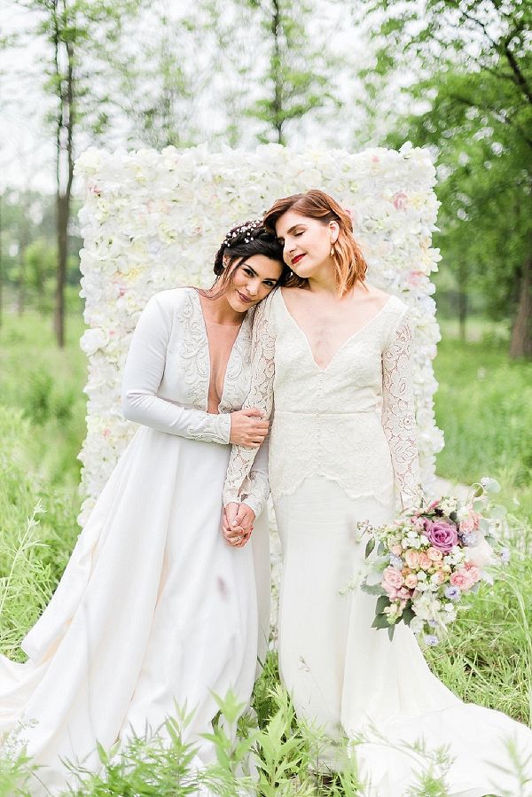 two brides pose for a photo in front of a flower covered backdrop at their wedding