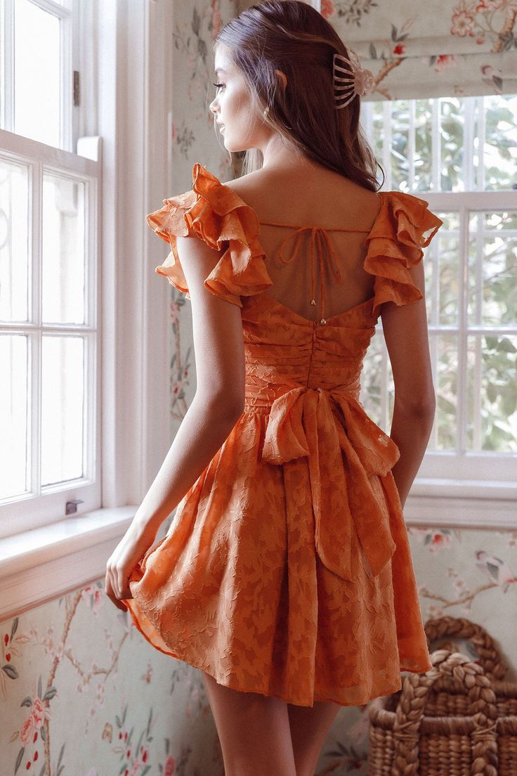 Orange textured dress Ruching detail in bodice Surplice neckline Ruffle shoulders Wide waistband Waist tie Scooped back with string tie Zipper in back You'll be the belle of the ball in our gorgeous Lola dress. We are in love with the ruching detail throughout the bodice and its flirty, layered ruffle shoulders. Perfect for a special birthday celebration or a garden wedding teamed with perspex heels and a clutch. Cute! MODEL INFO Model is wearing size XS Height: 5'8" Bust: 31" Waist: 23.5" Hips: Ruffle Homecoming Dresses, Orange Spring Dress, Short Colorful Dress, Fall Orange Dress, Peach Orange Dress, Orange Aesthetic Dress, Orange Graduation Dress, Orange Dress Aesthetic, Sunset Orange Dress