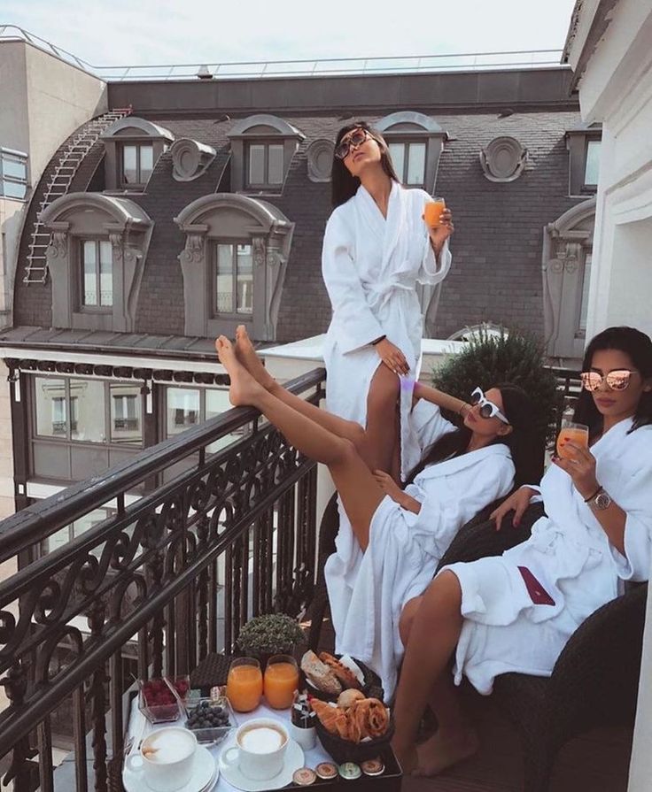 three women in white robes sitting on a balcony drinking orange juice and having breakfast together