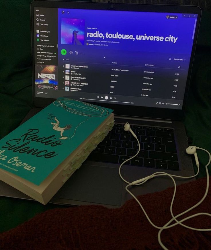 an open laptop computer sitting on top of a bed next to a book and headphones