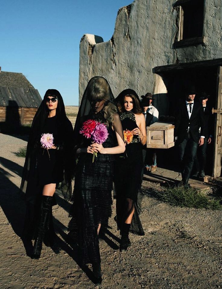 two women in black dresses walking down the street with people dressed up and one woman holding flowers