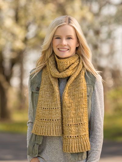 a woman wearing a yellow knitted scarf standing in front of a tree with her hands in her pockets