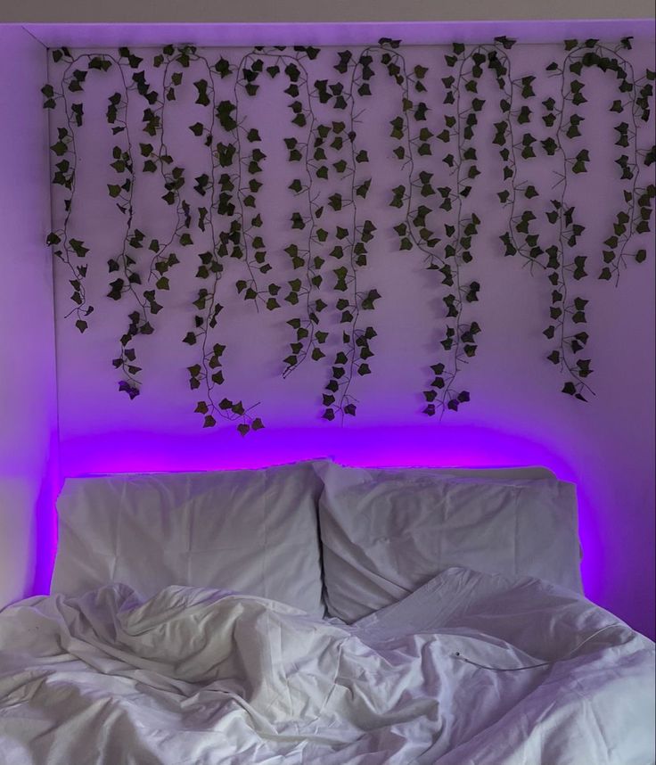 an unmade bed with white sheets and purple lights on the headboard is in front of a plant covered wall