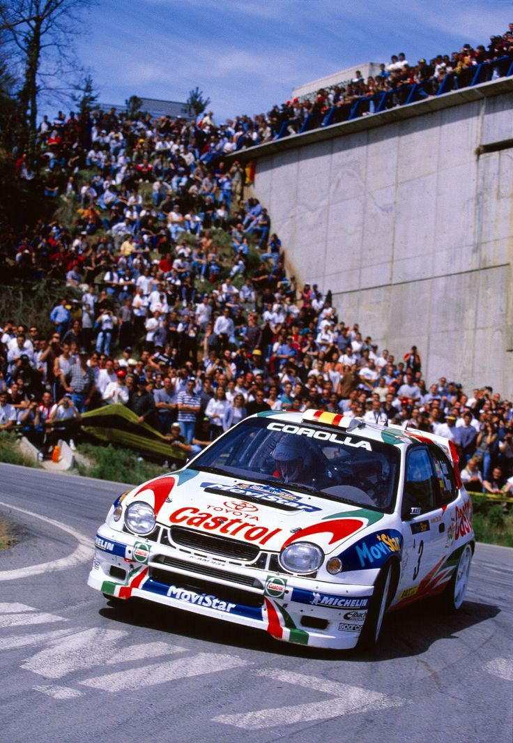 a rally car driving down a race track in front of a large crowd onlookers