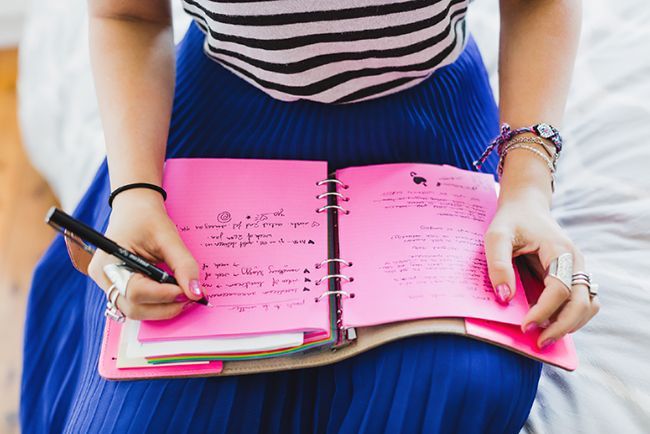 a woman holding a pink notebook and pen in her hand while sitting on a bed