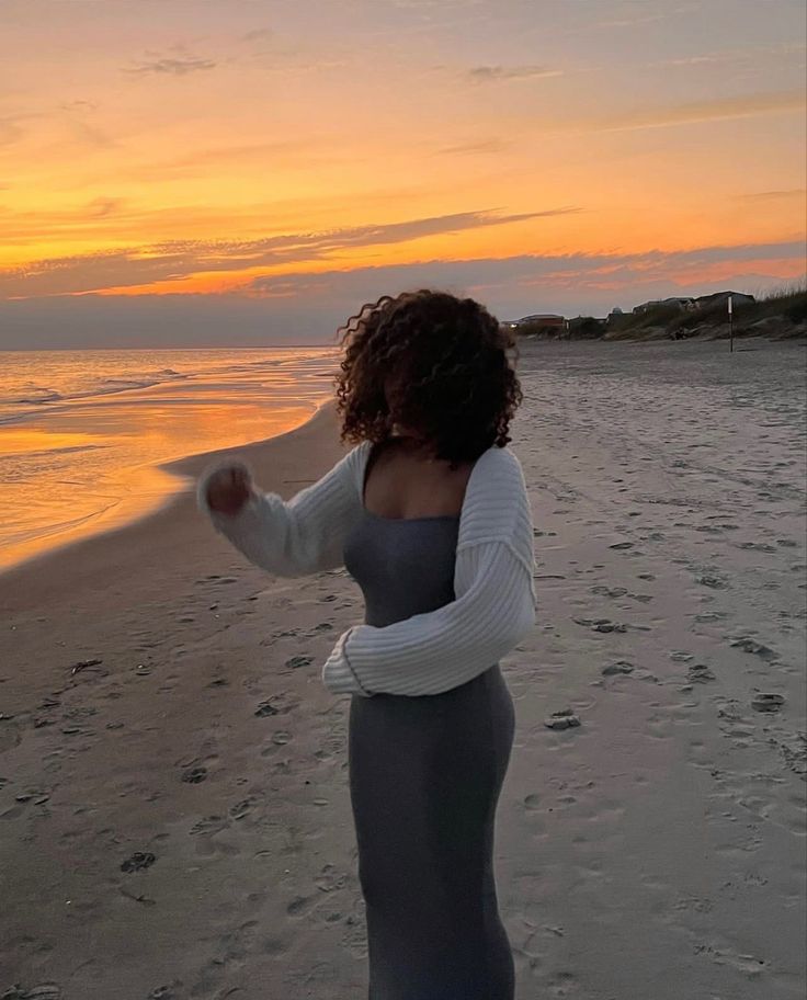 a woman standing on top of a sandy beach next to the ocean at sun set