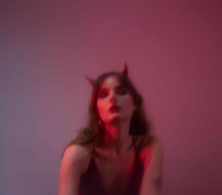 a woman with horns on her head sitting in front of a pink wall and looking at the camera