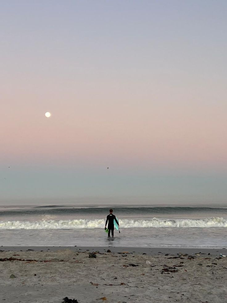 a person holding a surfboard on top of a sandy beach near the ocean at sunset