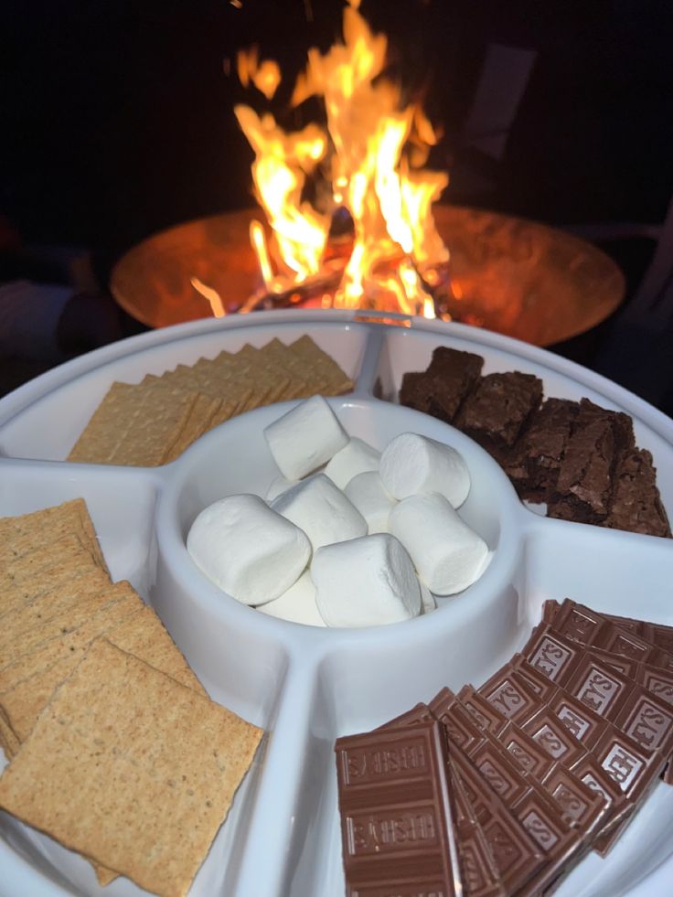 a plate with marshmallows, chocolate and graham crackers on it in front of a fire