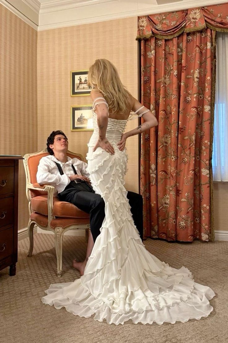 a woman in a white wedding dress standing next to a man sitting on a chair