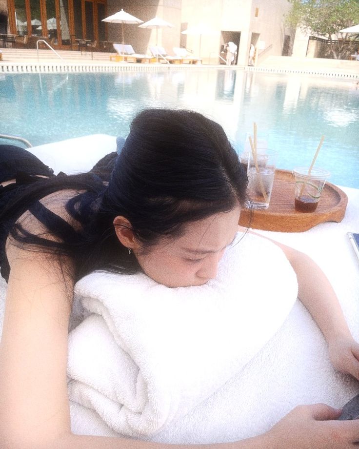 a woman laying on top of a towel next to a pool holding a cell phone