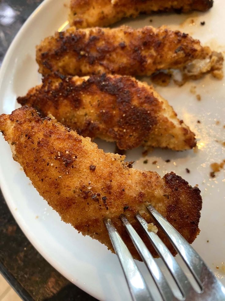 two pieces of chicken on a white plate with a fork in the foreground, and another piece of meat that has been fried