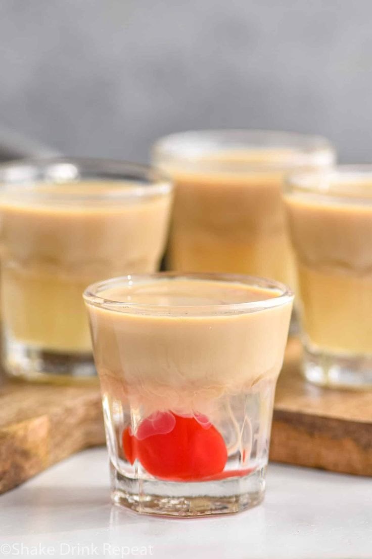 three shot glasses filled with liquid and a cherry on the rim, sitting on a cutting board