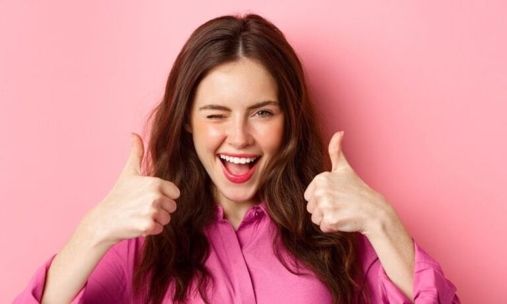 a woman in a pink shirt is giving the thumbs up sign with both hands while standing against a pink background