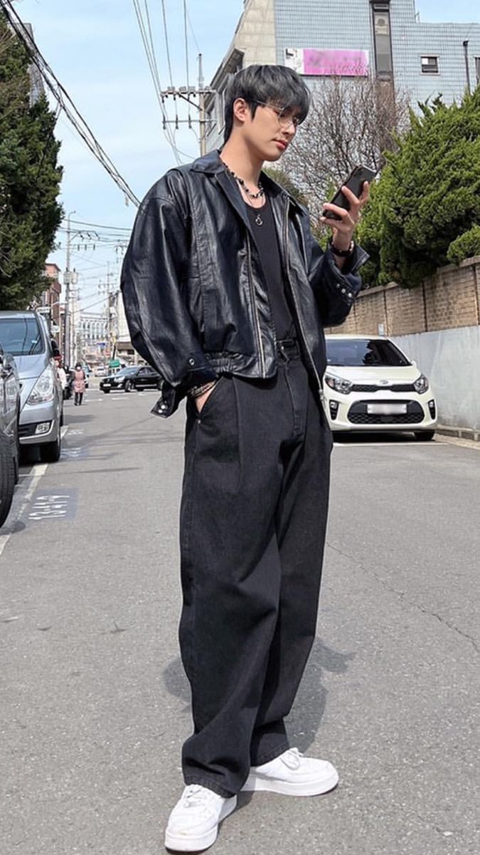 a man standing on the street looking at his cell phone while wearing black pants and a leather jacket