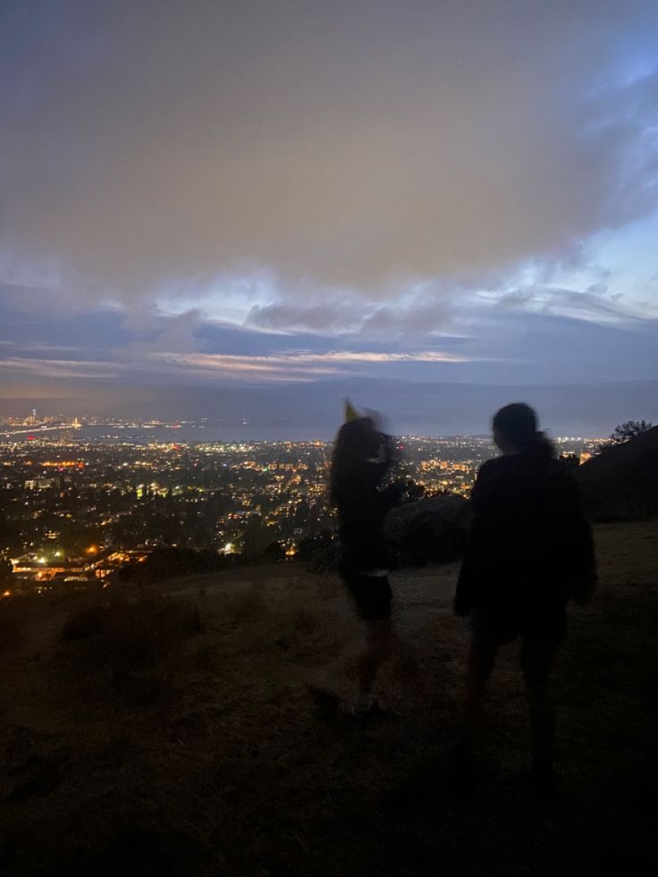 two people standing on top of a hill looking at the city lights in the distance