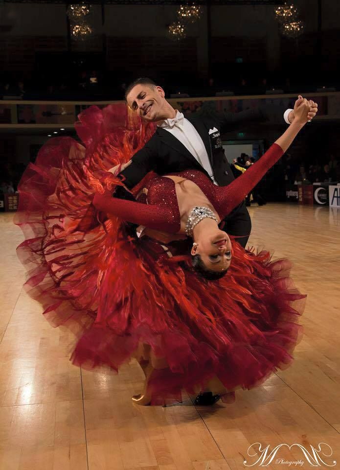 a man and woman in red dress dancing on a wooden floor with chandeliers hanging from the ceiling