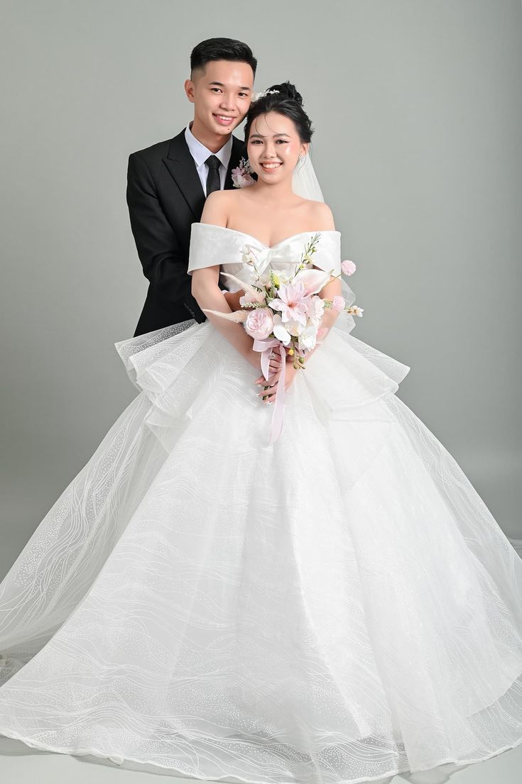 a bride and groom posing for a photo in front of a gray background wearing wedding gowns