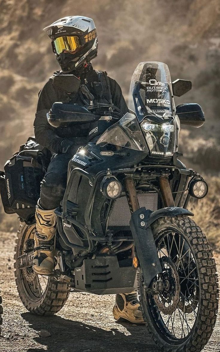 a man riding on the back of a black motorcycle down a dirt road next to a mountain