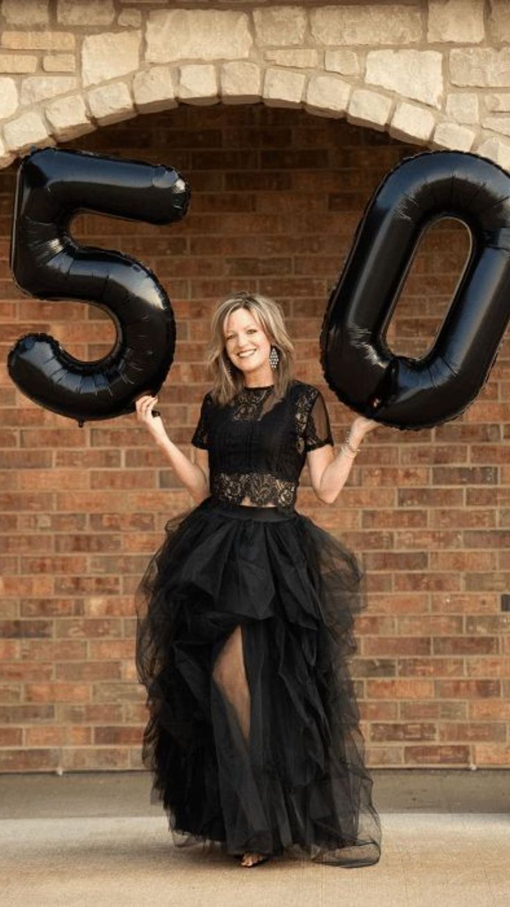 a woman standing in front of a brick wall holding two large black balloons that spell the number 50