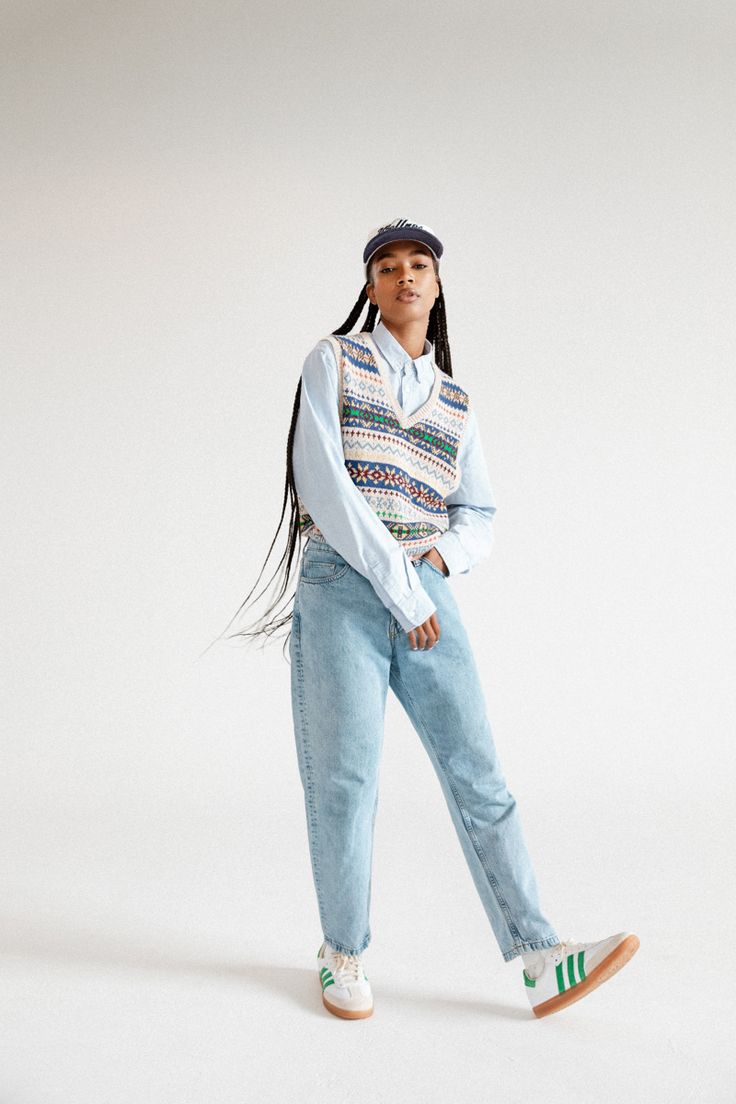 a woman standing in front of a white background wearing blue jeans and a colorful sweater