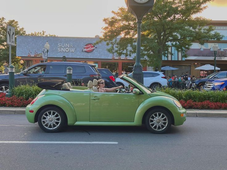 a woman driving a green convertible car down a street in front of a clock tower