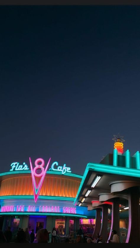 the outside of a restaurant at night with neon lights on it's roof and people walking around