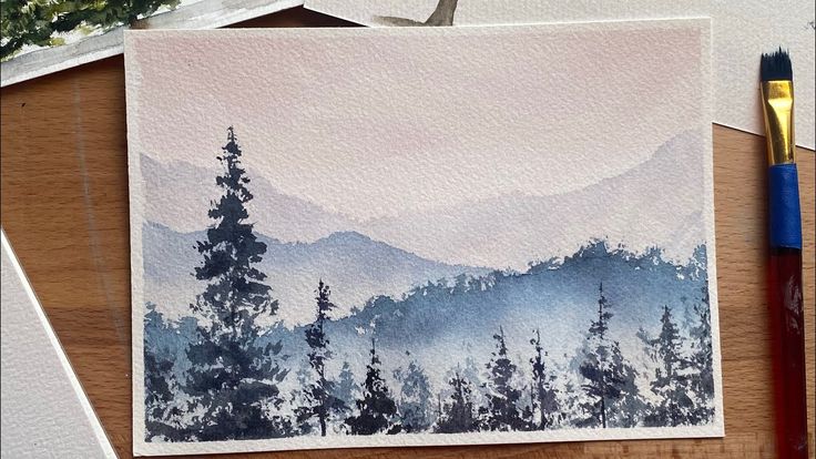 a watercolor painting of mountains and trees on a table with paintbrushes next to it