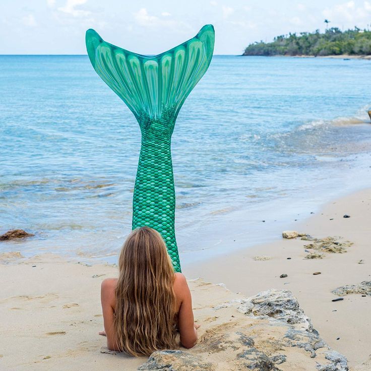 a woman laying on top of a sandy beach under a green mermaid tail sculpture next to the ocean