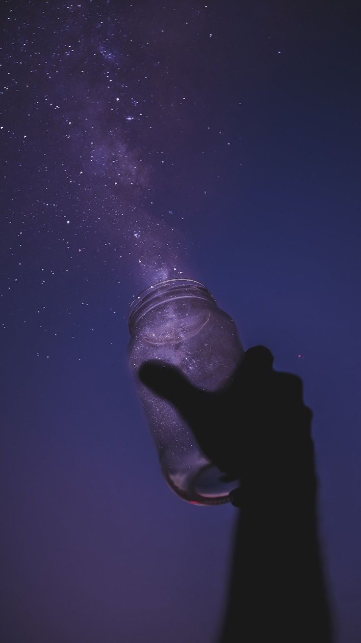 a hand holding a jar with stars in the sky above it and pointing at something