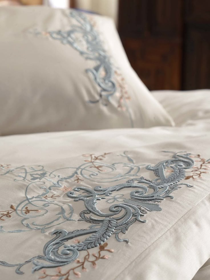 a close up of a bed with white sheets and blue embroidered designs on the pillows