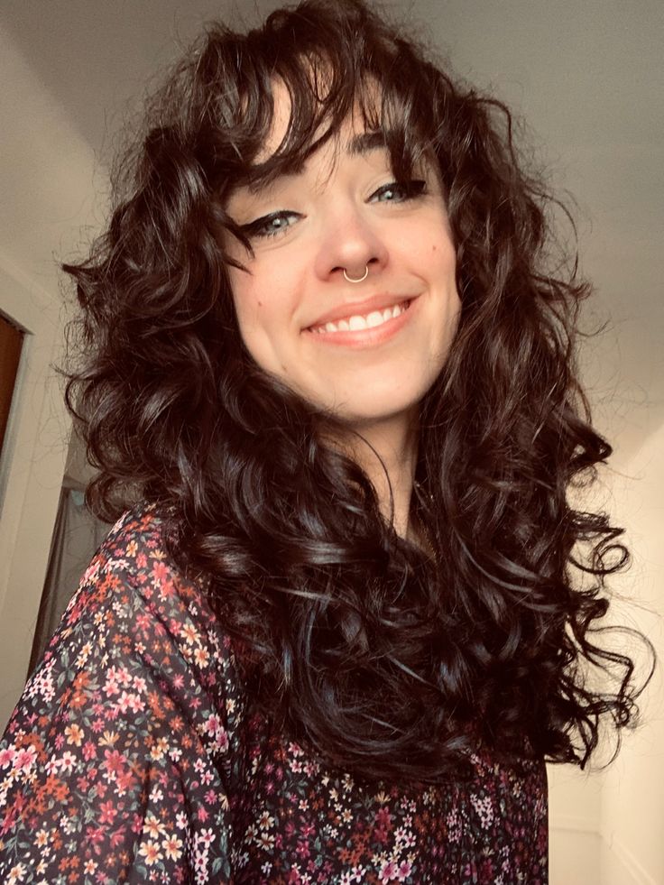 Girl smiling in selfie photo. Long brown curly hair with bangs, blue eyes with black winged eye-liner, septum ring in nose. Wearing a dark floral top. Med Length Curly Hair With Bangs, Butterfly Haircut With Bangs Curly Hair, Loose Curly Hair With Bangs, Layered Chunky Curls With Bangs, Shoulder Length 2b Hair, Medium Curly Shag With Bangs, Curly Layers With Bangs, Curly Shag Haircut With Bangs, Curly Hair Cuts Bangs