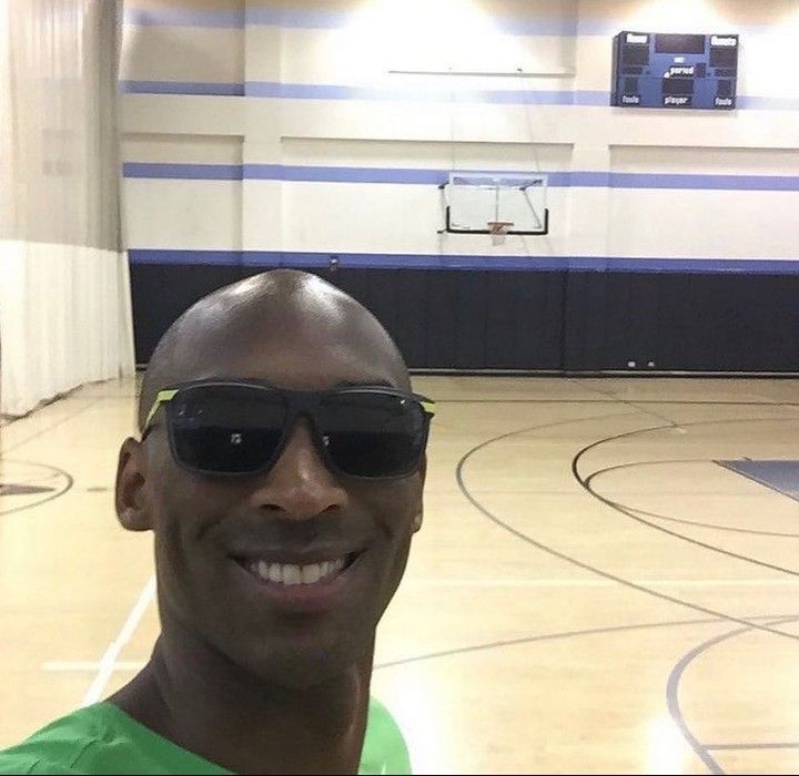 a man wearing sunglasses and smiling for the camera in a gym with basketball hoop behind him