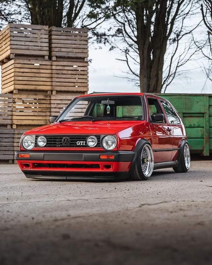 a red vw golf car parked in front of stacks of wooden pallet boxes