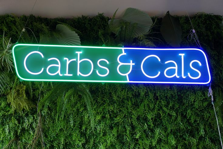 a neon sign that says cars and cats on it next to a wall of plants