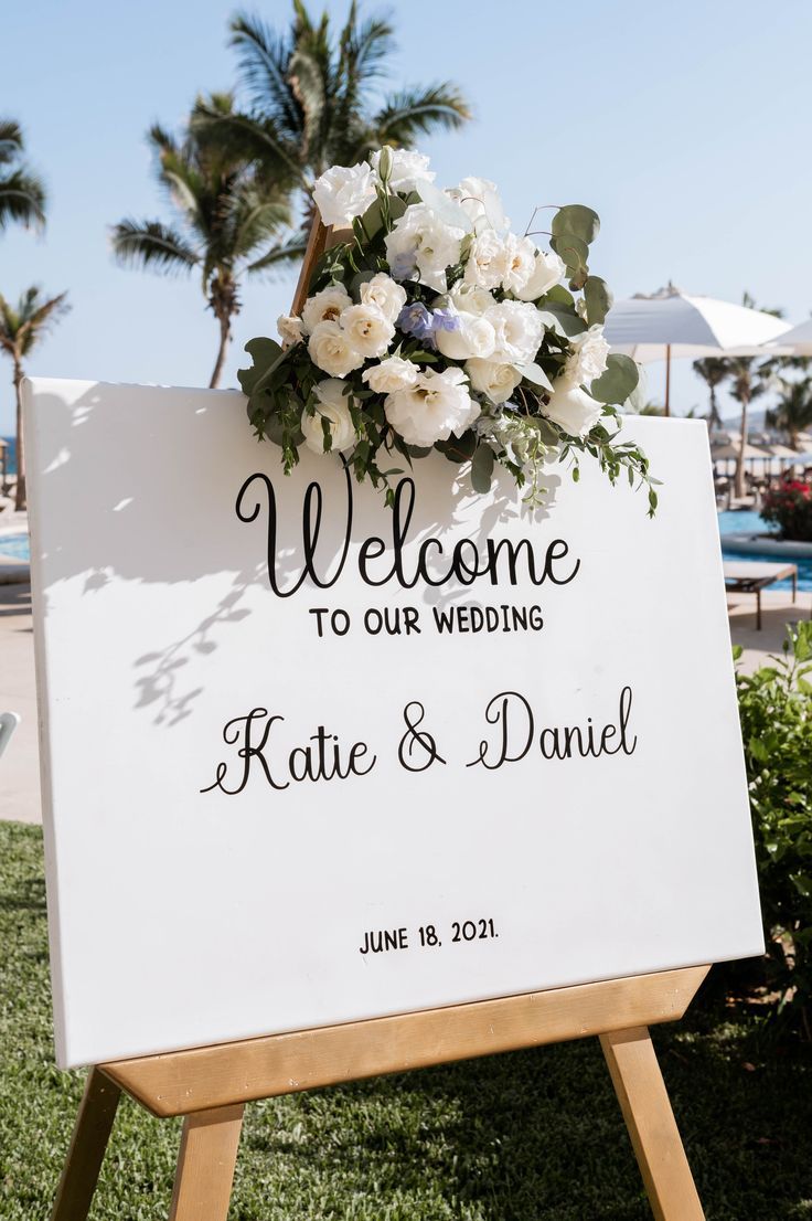 a welcome sign with flowers on it in front of some palm trees and lawn chairs